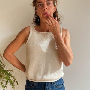 May include: A cream-coloured tank top with a square neckline and a ribbed texture. The top is paired with blue denim jeans. The model is wearing gold jewellery, including hoop earrings and a bracelet.