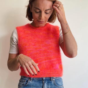 Pode incluir: Um colete de malha rosa e laranja difuso, usado sobre uma camiseta branca e jeans azul.