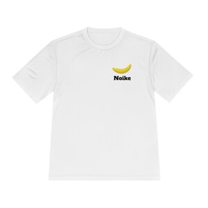May include: White t-shirt featuring a yellow banana graphic and the word "Noike" in black. The banana is slightly curved, with a light yellow color and a dark yellow outline. The t-shirt has short sleeves and a crew neck.