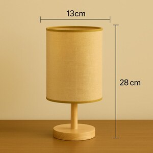 May include: A cylindrical table lamp with a beige fabric shade, 5 inches in diameter and 11 inches tall, sits on a wooden base. The lamp has a simple, minimalist design, suitable for a bedroom or living room.