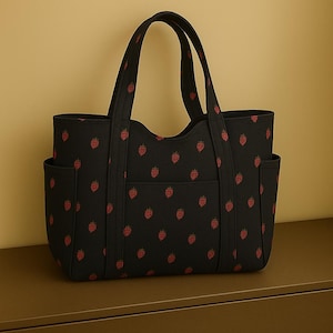 May include: Black tote bag featuring a repeating pattern of red strawberries. The bag has two external pockets and a front pocket. The handles are black. The bag is made of a durable material and is on a brown surface.