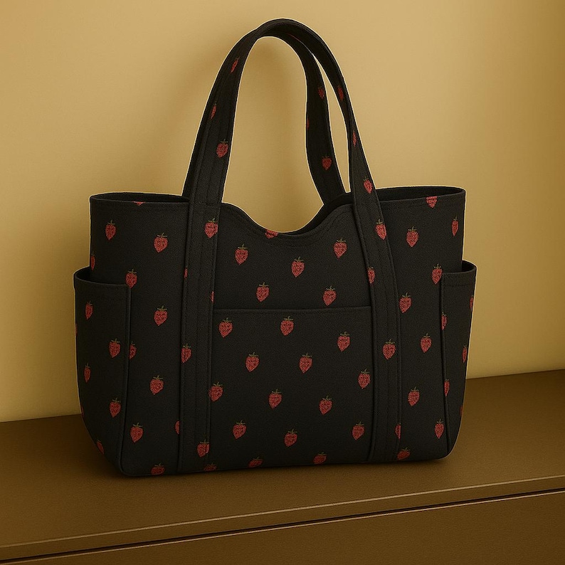 May include: Black tote bag featuring a repeating pattern of red strawberries. The bag has two external pockets and a front pocket. The handles are black. The bag is made of a durable material and is on a brown surface.