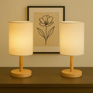 Pode incluir: Duas luminárias de mesa combinando com abajures cilíndricos brancos e bases de madeira. As luminárias estão em uma superfície de madeira escura, com uma impressão floral emoldurada no fundo. As luminárias fornecem uma iluminação suave e quente.