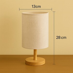 May include: A small table lamp with a beige fabric shade, 5 inches in diameter and 11 inches tall, sits on a wooden base. The lamp has a cylindrical shade and a wooden stem. The lamp is designed for indoor use.
