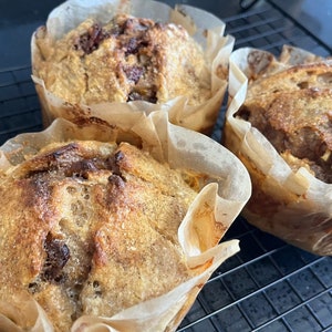 May include: Three golden brown muffins in paper liners, sitting on a black wire rack. The muffins have a slightly crispy top and are slightly domed in shape.