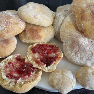 May include: An assortment of freshly baked bread rolls and flatbreads, some with a golden-brown crust. Two rolls are cut open and spread with butter and a vibrant red jam. The bread is arranged on a white surface.