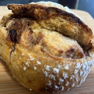 May include: A close-up of a round, brown bread roll with a swirl of cinnamon and raisins. The bread is dusted with a light layer of powdered sugar.