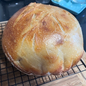 May include: A freshly baked loaf of sourdough bread with a golden brown crust. The bread has a slightly domed shape and is sitting on a wire rack.