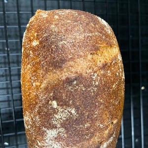 May include: A loaf of freshly baked bread with a golden-brown crust, resting on a black wire rack. The bread has a rustic appearance with a textured surface and a crack along the top. The color is a mix of brown and tan.