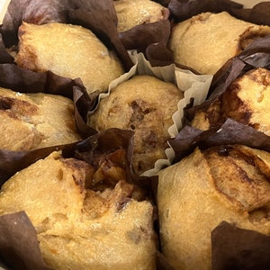 May include: Close-up of six brown cinnamon rolls in individual paper liners, arranged in a circle in a metal baking pan. The rolls are golden brown and have a slightly crispy crust.