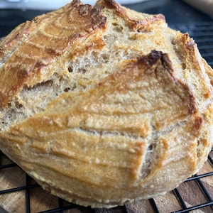 May include: A freshly baked loaf of sourdough bread with a golden brown crust. The bread has a slightly irregular shape and a textured surface.