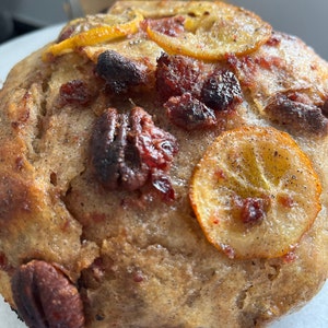 May include: A close-up of a baked good with a brown crust, topped with dried cranberries, pecans, and lemon slices. The baked good has a slightly glossy finish.