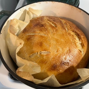 以下が含まれることがあります： 鋳鉄製のダッチオーブンに入った、焼き立てのパン。パンは黄金色で、カリカリのクラスト。パンはクッキングシートの上に置かれています。