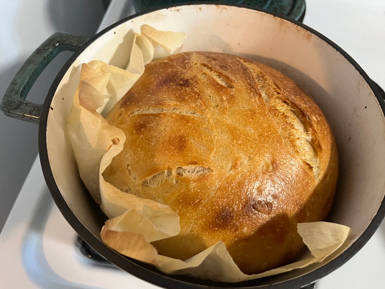 以下が含まれることがあります： 鋳鉄製のダッチオーブンに入った、焼き立てのパン。パンは黄金色で、カリカリのクラスト。パンはクッキングシートの上に置かれています。