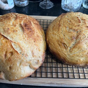 以下が含まれることがあります： ワイヤーラックの上に置かれた、焼き立てのパン2つが、木のまな板の上で冷まされています。パンは、黄金色の焼き色がついており、形が少し不揃いです。
