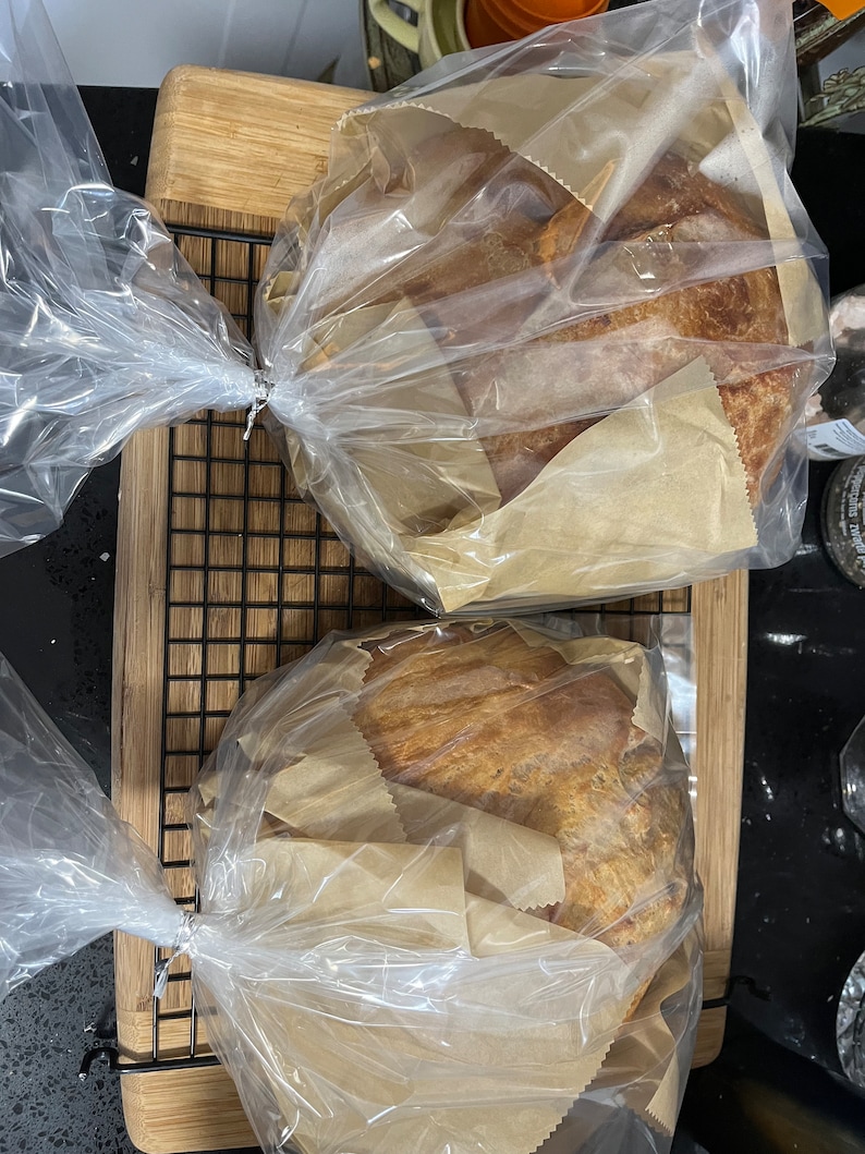 以下が含まれることがあります： 2つのパンが茶色の紙と透明なビニール袋に包まれています。パンは木製の表面の黒いワイヤーラックの上にあります。