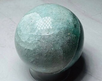 Puede incluir: Una esfera pulida de cristal de aventurina, verde y blanca, sobre un soporte negro.