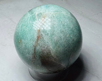 Puede incluir: Una esfera verde y blanca con vetas marrones y blancas, sentada sobre un soporte negro. La esfera está hecha de piedra pulida.