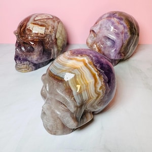May include: Three amethyst skull sculptures, each with unique banding and color variations. The skulls are carved from natural amethyst and have a polished finish.