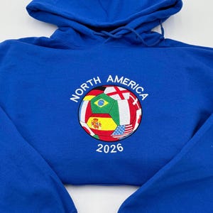 May include: Royal blue hoodie with a drawstring hood. The front features a soccer ball design with flags of various nations and the text "NORTH AMERICA 2026". The design is embroidered in multiple colors.