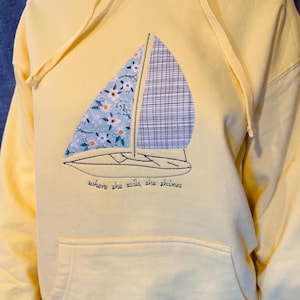 May include: A yellow hooded sweatshirt featuring an embroidered sailboat design. The sailboat has a floral patterned sail and a plaid patterned sail. The text "where she sails she shines" is embroidered below the sailboat.