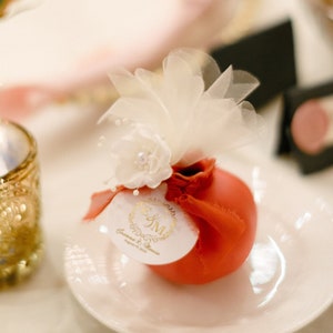 May include: A small red favor box with a white tulle bow and a white circle sticker with gold lettering that says "Save the Date" and "E&M" in a monogram. The box is on a white plate.