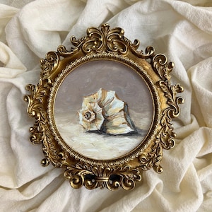 Op de afbeelding: Schilderij van een zeeschelp in een sierlijke gouden lijst. Het kunstwerk toont een zeeschelp in tinten wit, beige en grijs. De lijst is rond en decoratief. De tekst "TuanArt Studio" is onderaan de afbeelding zichtbaar.