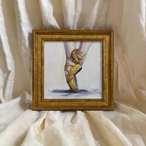 Op de afbeelding: Een ingelijst schilderij van balletschoenen in een gouden lijst. Het schilderij toont de schoenen in een gouden tint, met de linten om de enkels gewikkeld. De achtergrond is een zachte, neutrale kleur. De initialen van de kunstenaar zijn zichtbaar.