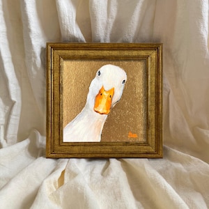 May include: A square painting of a white duck with an orange beak set against a metallic gold background, enclosed in a textured gold frame. The artwork features a small orange signature in the bottom right corner of the painted surface.
