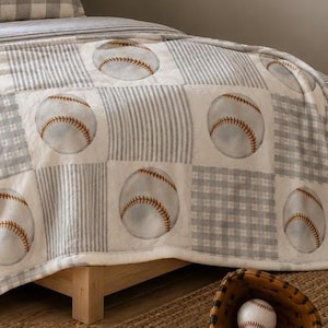 Puede incluir: Una manta suave con un patrón repetitivo de pelotas de béisbol sobre cuadrados alternos de cuadros vichy grises y blancos y rayas. Una almohada rectangular con el número 07 en azul marino descansa sobre la cama, junto a un guante de béisbol de cuero marrón.