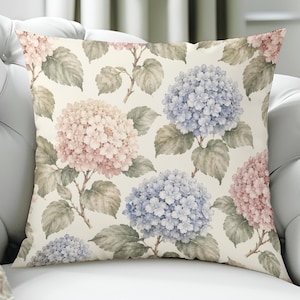 Könnte beinhalten: Ein quadratisches Zierkissen mit einem sich wiederholenden botanischen Muster aus Hortensienblüten in zarten Pastellrosa- und Hellblautönen, akzentuiert durch gedämpfte grüne Blätter auf einem cremeweißen Hintergrund mit Blumendruck.