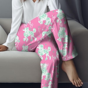 Op de afbeelding: Roze pyjamabroek met een wit silhouetpatroon van poedels. De poedels zijn omlijnd met een lichtblauwe kleur en hebben een strik aan hun staart.