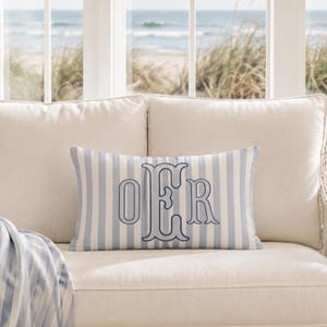 May include: A rectangular throw pillow featuring a light blue and white vertical striped pattern. Centered on the pillow is a large, dark blue embroidered monogram consisting of the letters O, E, and R in a decorative, serif-style font.