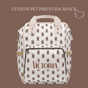 Puede incluir: Mochila blanca con un patrón repetido de caras de perro. El bolsillo delantero muestra el nombre "Victoria" en marrón. La mochila tiene un asa y correas ajustables. El texto "CUSTOM PET PHOTO BACKPACK" está en la parte superior.