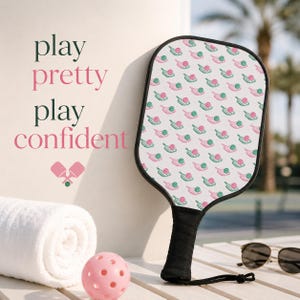 Könnte beinhalten: Ein Pickleball-Schläger mit schwarzem Griff und einer gemusterten Oberfläche mit rosa und grünen Pickleball-Grafiken. Ein rosa Pickleball, ein weißes Handtuch und eine Sonnenbrille sind ebenfalls sichtbar. Der Text lautet "play pretty play confident."