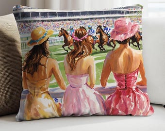 May include: Decorative throw pillow featuring a watercolor painting of three women watching a horse race. The women are wearing dresses and hats in shades of yellow, pink, and red. The background shows the race track and a crowd of spectators.