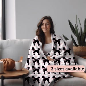 Op de afbeelding: Een wit fleece deken met een zwart poedel silhouetpatroon en roze strikken. Het deken is over een bank gelegd en een persoon zit op de bank. De tekst "3 maten beschikbaar" staat in de rechterbenedenhoek van de afbeelding.