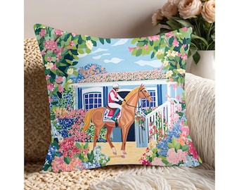 May include: Decorative throw pillow featuring a horse and jockey in front of a white building with blue shutters. The scene is surrounded by colorful flowers in shades of pink, blue, and green. The pillow is on a cream-colored textured surface.