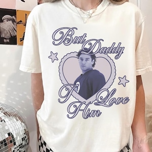 Puede incluir: Camiseta color crema con un gráfico morado en forma de corazón. El diseño presenta un retrato de un hombre y el texto "But Daddy I Love Him" en una fuente decorativa, con estrellas.