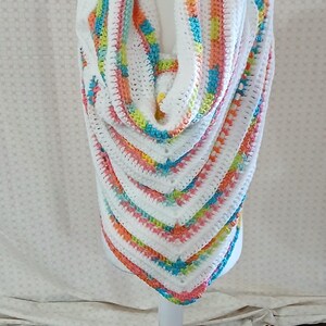 May include: A white crochet shawl with colorful stripes of orange, yellow, blue, and pink. The shawl is displayed on a white mannequin. The shawl is triangular in shape and appears to be handmade.