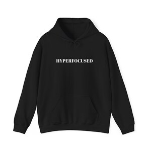 Op de afbeelding: Zwart hoodie met de tekst "HYPERFOCUSED" in wit gedrukt op de voorkant.