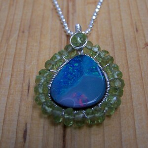 Pu&ograve; includere: Una collana con catena d'argento con un ciondolo a forma di lacrima che presenta una pietra di opale blu e verde circondata da perline di peridoto verdi.
