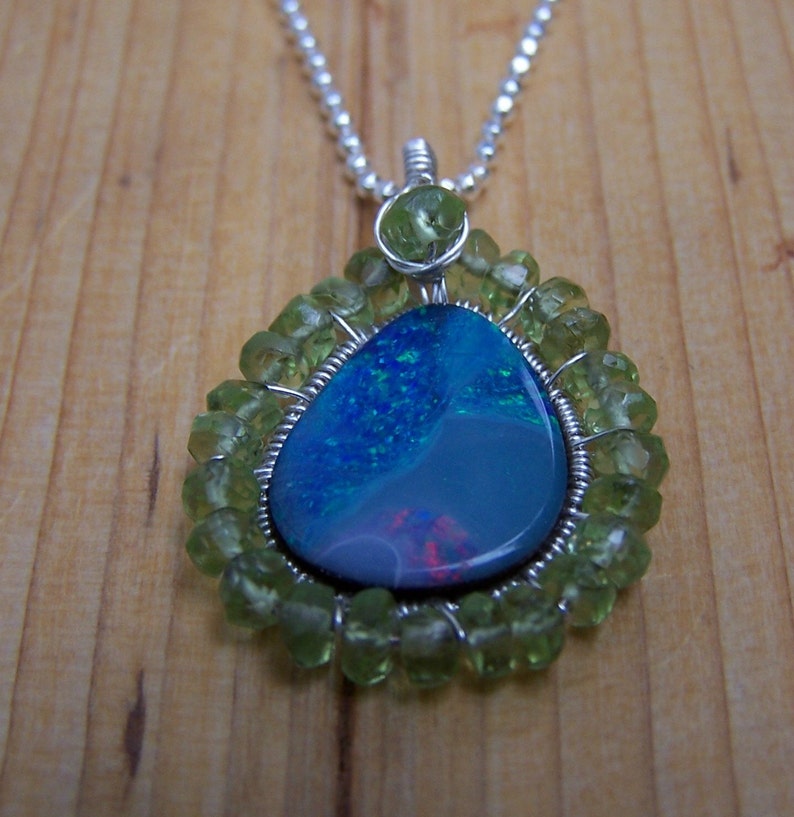 Pu&ograve; includere: Una collana con catena d'argento con un ciondolo a forma di lacrima che presenta una pietra di opale blu e verde circondata da perline di peridoto verdi.