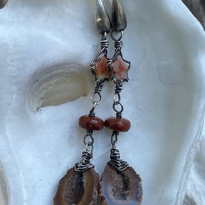 May include: A pair of dangle earrings featuring silver-toned wire-wrapped star-shaped orange stones, round dark red beads, and bottom pendants consisting of brown geode slices with crystalline centers, suspended from silver-toned metal hooks.