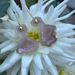 Puede incluir: Un par de pendientes rosa claro y dorados con una forma orgánica única. Los pendientes tienen un poste dorado y una piedra texturizada rosa claro. Los pendientes se muestran sobre una flor blanca con centro amarillo.