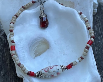 May include: A beaded bracelet featuring a central white ceramic bead with a red and blue floral pattern, flanked by silver-toned metal spacers. The strand includes small beige and bright red beads on a dark cord, finished with a dark red teardrop charm.