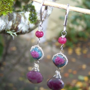 May include: A pair of silver earrings with pink and blue gemstones. The earrings feature a teardrop-shaped pink gemstone, a round blue gemstone, and a small pink bead.