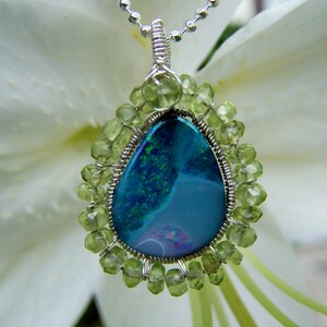 Pu&ograve; includere: Una collana con catena d'argento con un ciondolo a forma di lacrima che presenta una pietra di opale blu e verde circondata da un bordo avvolto in filo di pietre preziose di peridoto verde.