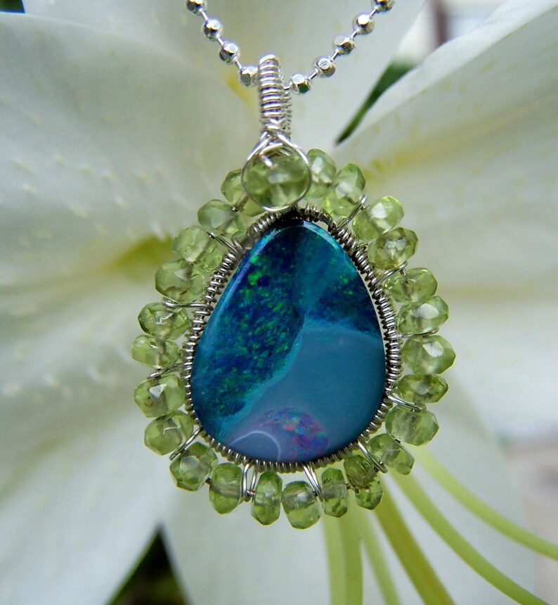 Pu&ograve; includere: Una collana con catena d'argento con un ciondolo a forma di lacrima che presenta una pietra di opale blu e verde circondata da un bordo avvolto in filo di pietre preziose di peridoto verde.