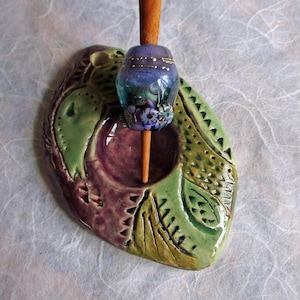 May include: A handcrafted ceramic spindle whorl in shades of green and purple, with a wooden spindle and a decorative glass bead. The bead features blue and purple floral designs and gold accents. The whorl is approximately 10 cm long.